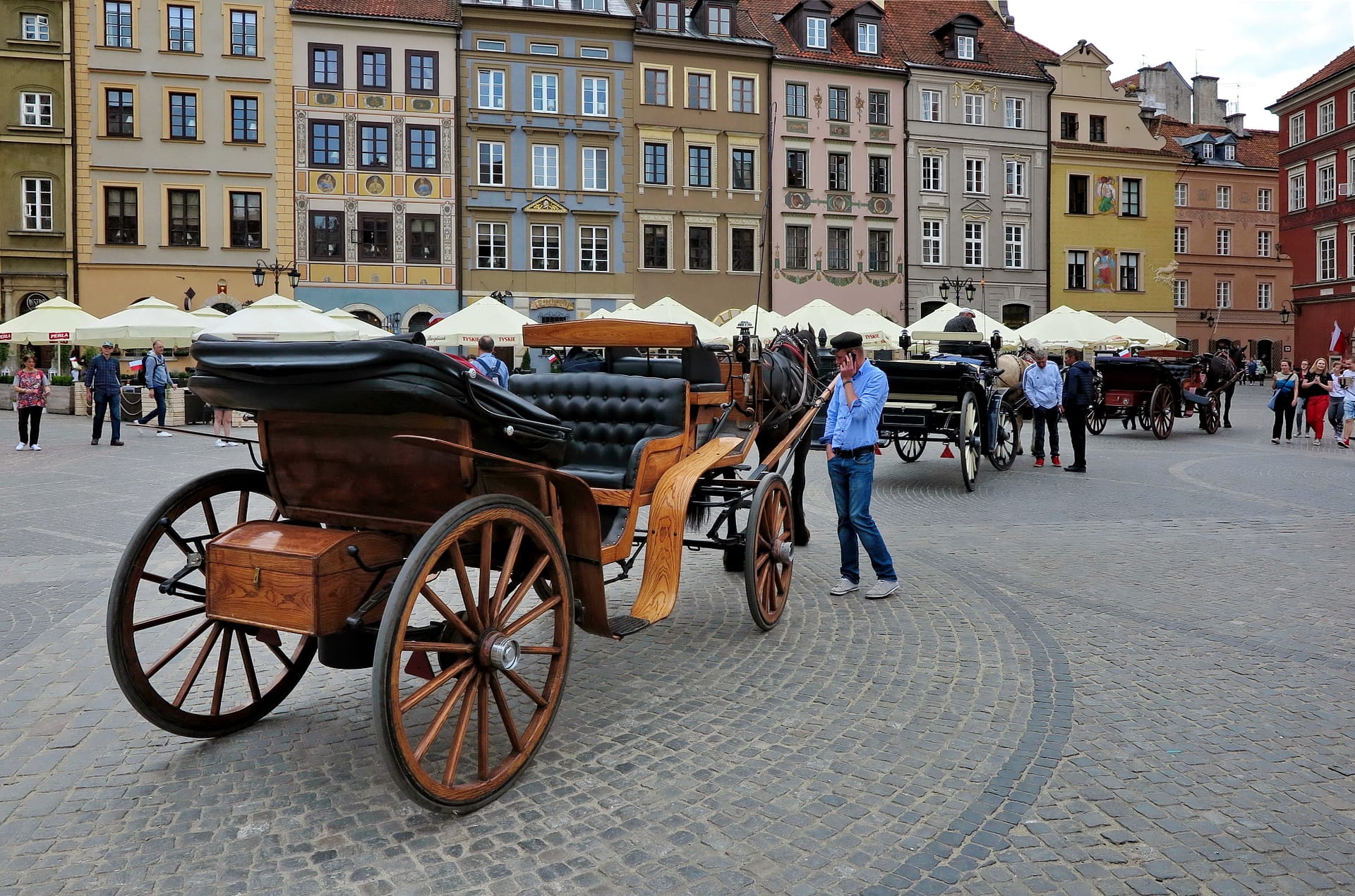 Old horse-drawn carriages await tourists at the old town Market square in the centre of Warsaw. The driver is talking on the phone. Old houses behind. Poland, may 2018
